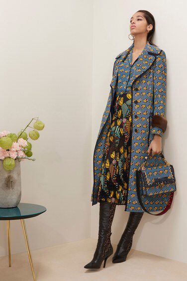 a fulllength shot of a fashion model posing in a stylish blue and yellow patterned coat, a black and yellow floral skirt, and black highheeled boots. she's carrying a matching handbag. the background is a light beige wall, and there's a small side table with flowers in a vase in the bottom left corner. the overall style is elegant and chic, suggesting an autumn or winter collection.
