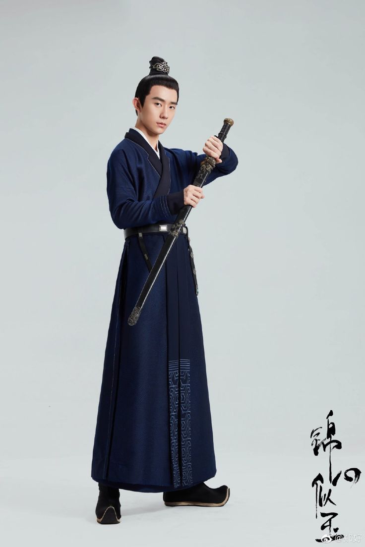 a full shot of a young asian man dressed in a traditional dark blue hanfu robe, holding a sword. he is standing against a plain light gray background, and his expression is serious. the image appears to be a promotional still for a film or television series. the costume is detailed, and the man's pose is elegant.  chinese calligraphy is visible in the bottom right corner.