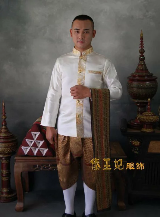 a thai man wearing traditional thai clothing. he is wearing a white longsleeve shirt with gold trim, brown pants with gold trim, and a brown sash. he is standing in front of a table with a traditional thai vase on it.
