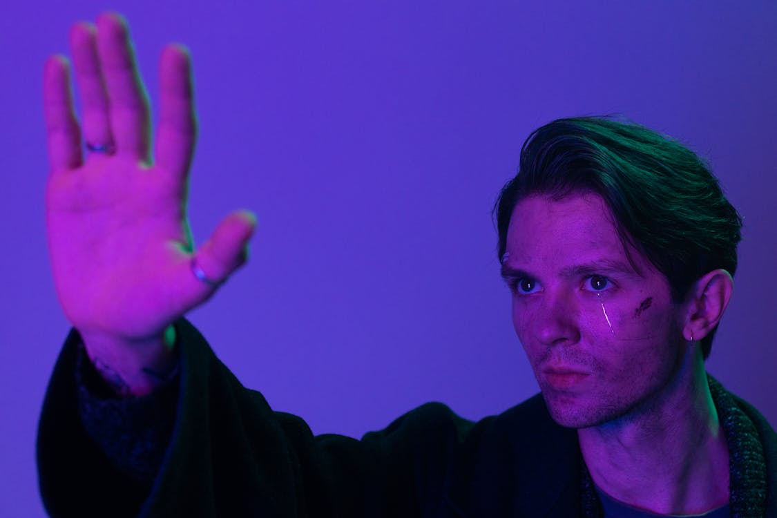 closeup portrait of a man with dark hair styled back, wearing a dark jacket. he is shown from the chest up, his left hand raised in a gesture that appears to be stopping something. the background is a solid purple with varying tones and intensities of purple and neon lighting.  his face shows an intense expression and he has a small mark or scar near his eye. the overall mood is dramatic and mysterious. the image is professionally lit and photographed.