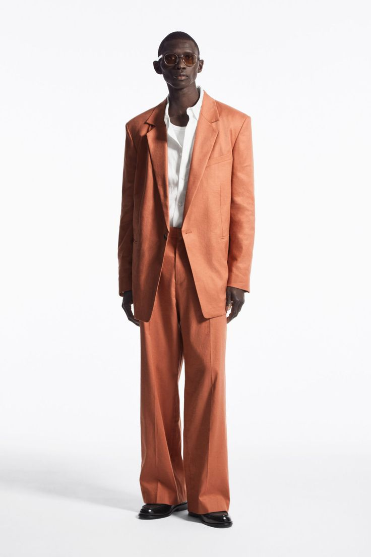 a man is wearing a orange suit with a white shirt and black sunglasses. he is standing in a studio with a white background and is looking at the camera. he is posing in a fashionable way. the suit is very stylish and the man looks confident and cool. this is a fashion editorial image.