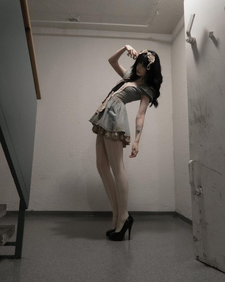 a woman in a dress, stockings, and heels is posing indoors in a staircase hallway. the walls are white, and there is a door to the right side. a gray staircase is on the left side. the woman has a tattoo on her arm and a headband in her hair.