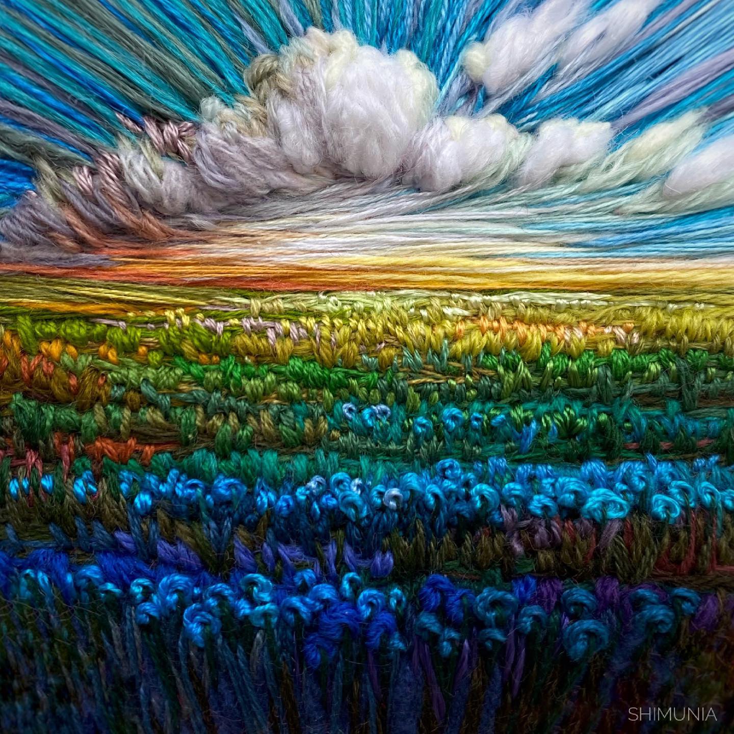 the image is a needlework art piece of a landscape. it is an embroidery created using various colored threads. the top of the image shows a bright sky with fluffy white and grey clouds.  the setting sun casts a warm glow across the horizon.  the bottom portion depicts a field of blue flowers with hints of green, yellow, orange and red.