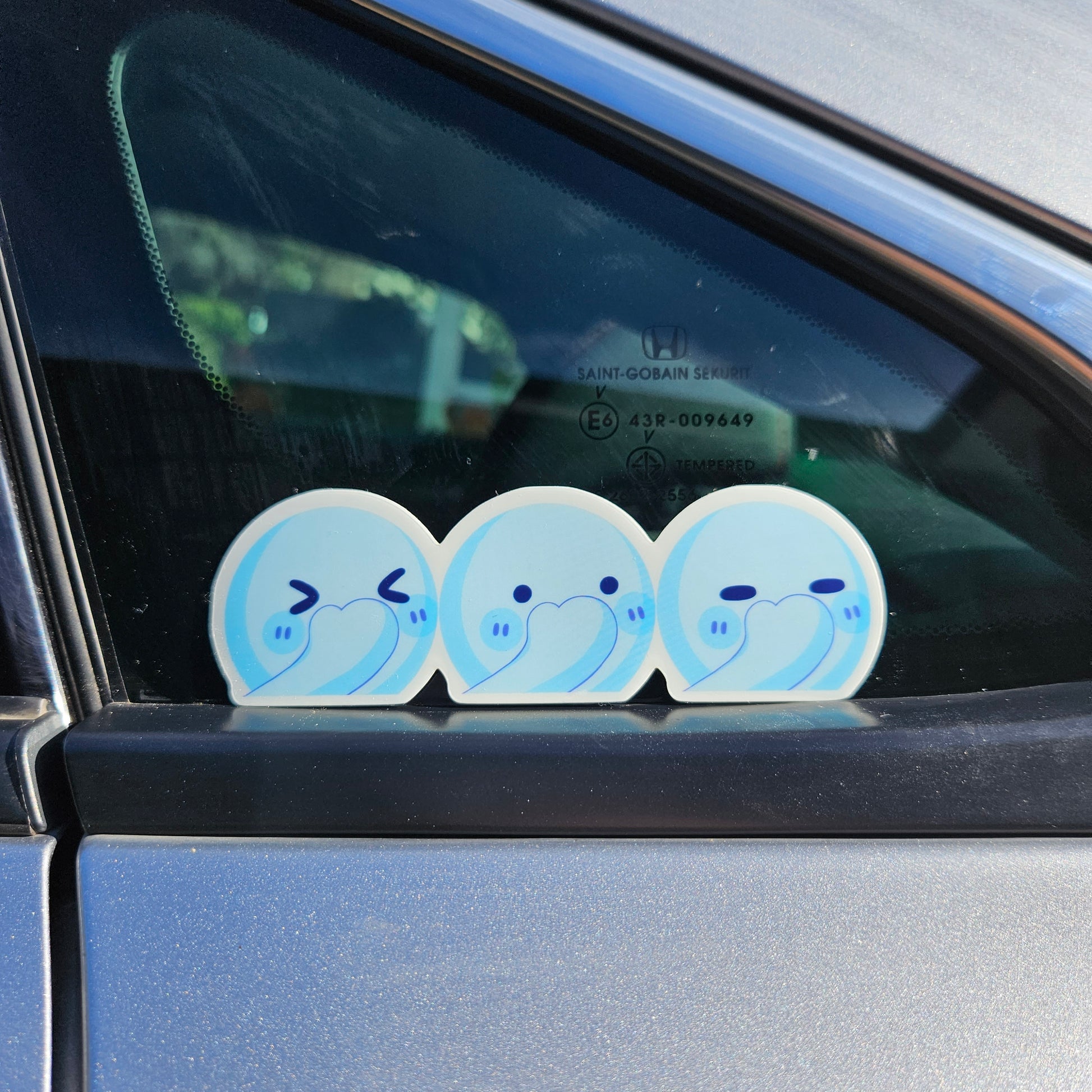 loseup view of three small, cute blue stickers applied to the rear passengerside window of a silver car. the stickers depict simple cartoon characters with shy or bashful expressions.  the window shows a partially visible reflection of the surrounding area, and a small portion of the car's safety glass markings are visible in the uppermiddle section of the image. the stickers are neatly arranged sidebyside. the overall impression is of a playful, whimsical addition to the vehicle.