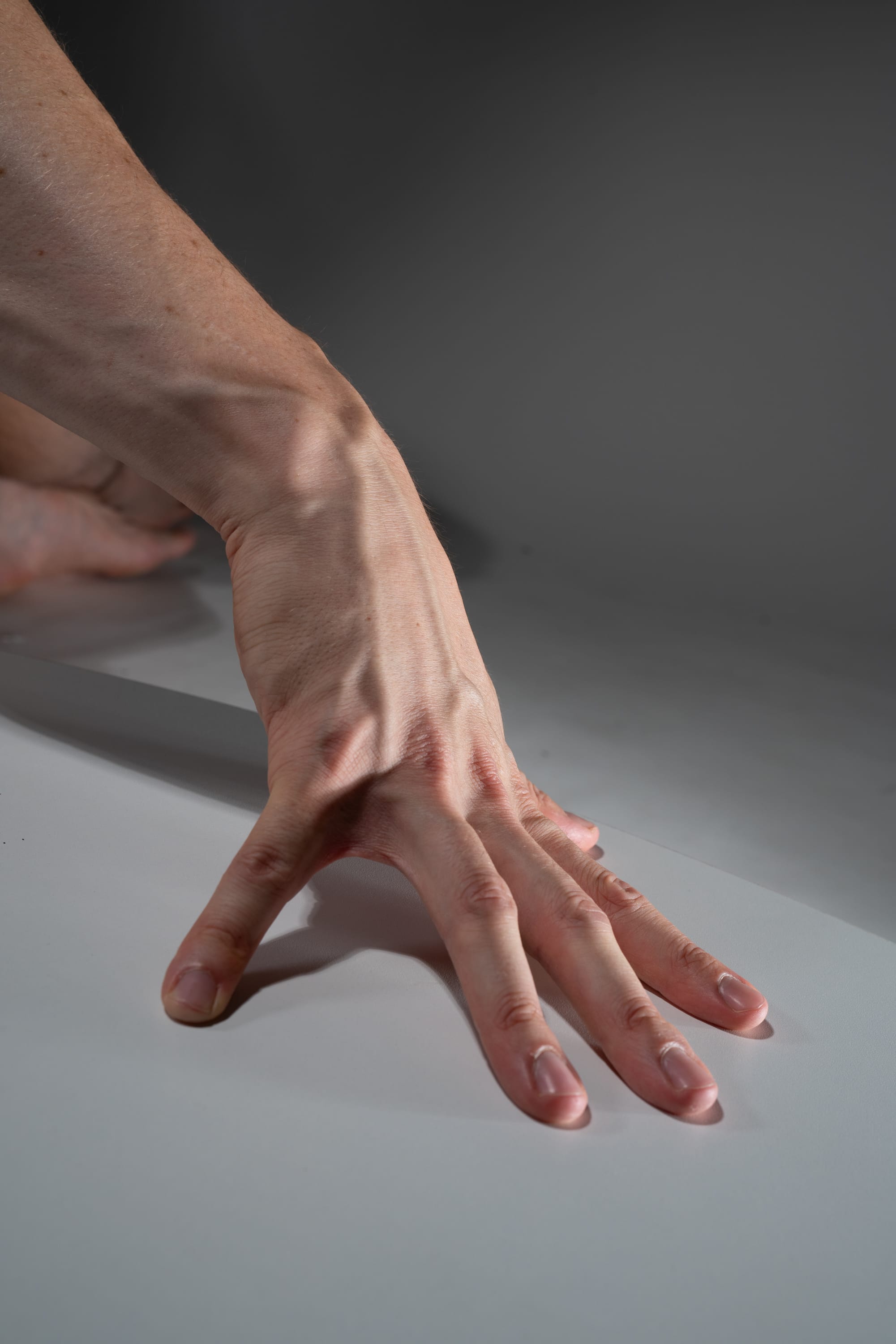 a closeup image of a hand with prominent veins and tendons, reaching out and pressing its fingers against a smooth, white surface. the hand is lit by studio lighting, highlighting its details and casting a subtle shadow beneath. the background is a muted grey, emphasizing the hand as the central focus.