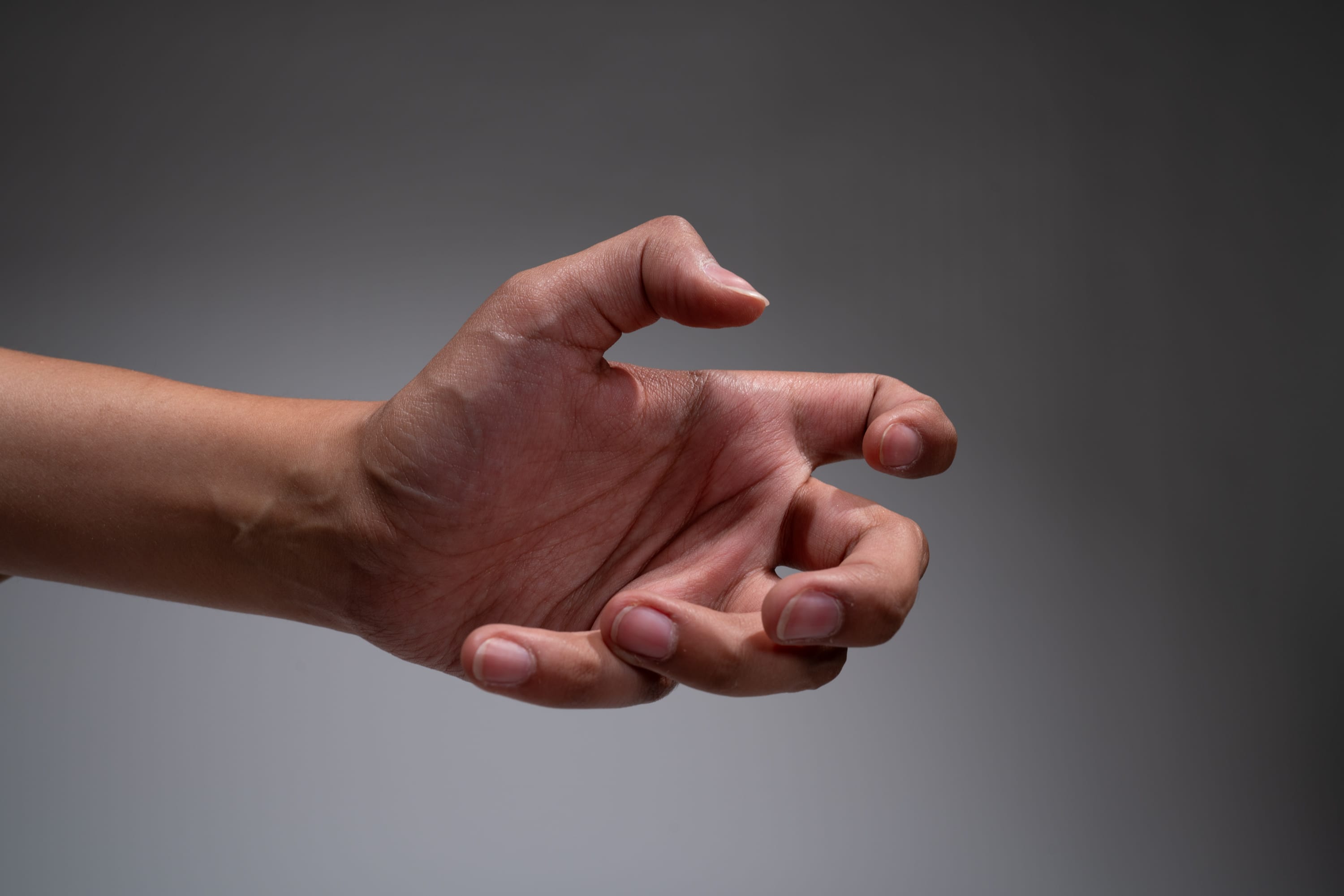 a hand is shown reaching out with fingers curled in a gesture of grabbing or holding something. the hand is positioned against a neutral gray background, illuminated by soft studio lighting. the image captures the hand from a side view at eye level, emphasizing the details of the skin and the gesture.