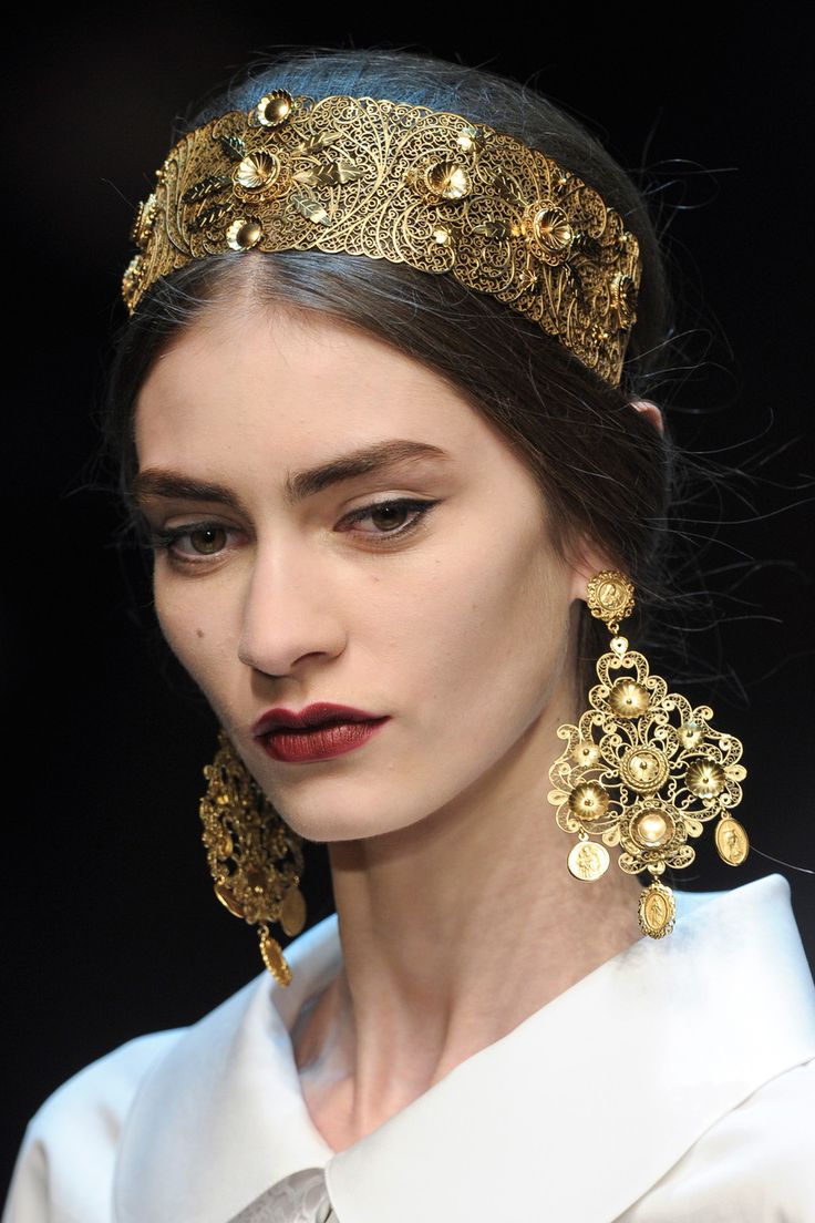loseup portrait of a fashion model wearing a stunning gold filigree headband and matching earrings. the jewelry is intricate and ornate, with a detailed design. the model has dark hair pulled back, dark lipstick, and is wearing a white collared shirt. the image is likely from a fashion runway show, showcasing highend, luxurious accessories. the overall aesthetic is elegant and regal.