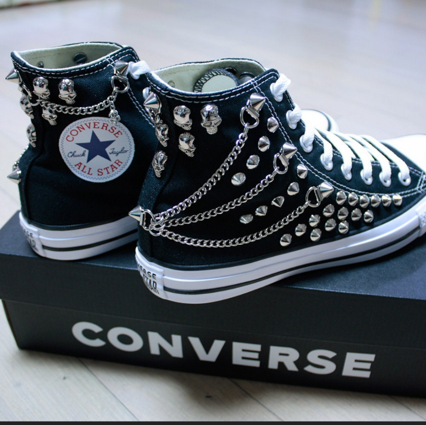 a closeup shot of a pair of customized black converse chuck taylor all star hightop sneakers.  the shoes are heavily decorated with silver metal studs, chains, and skull charms, giving them a punk rock or goth aesthetic. they are sitting in a converse shoebox.  the image is welllit and focuses on the detail of the modifications to the shoes.