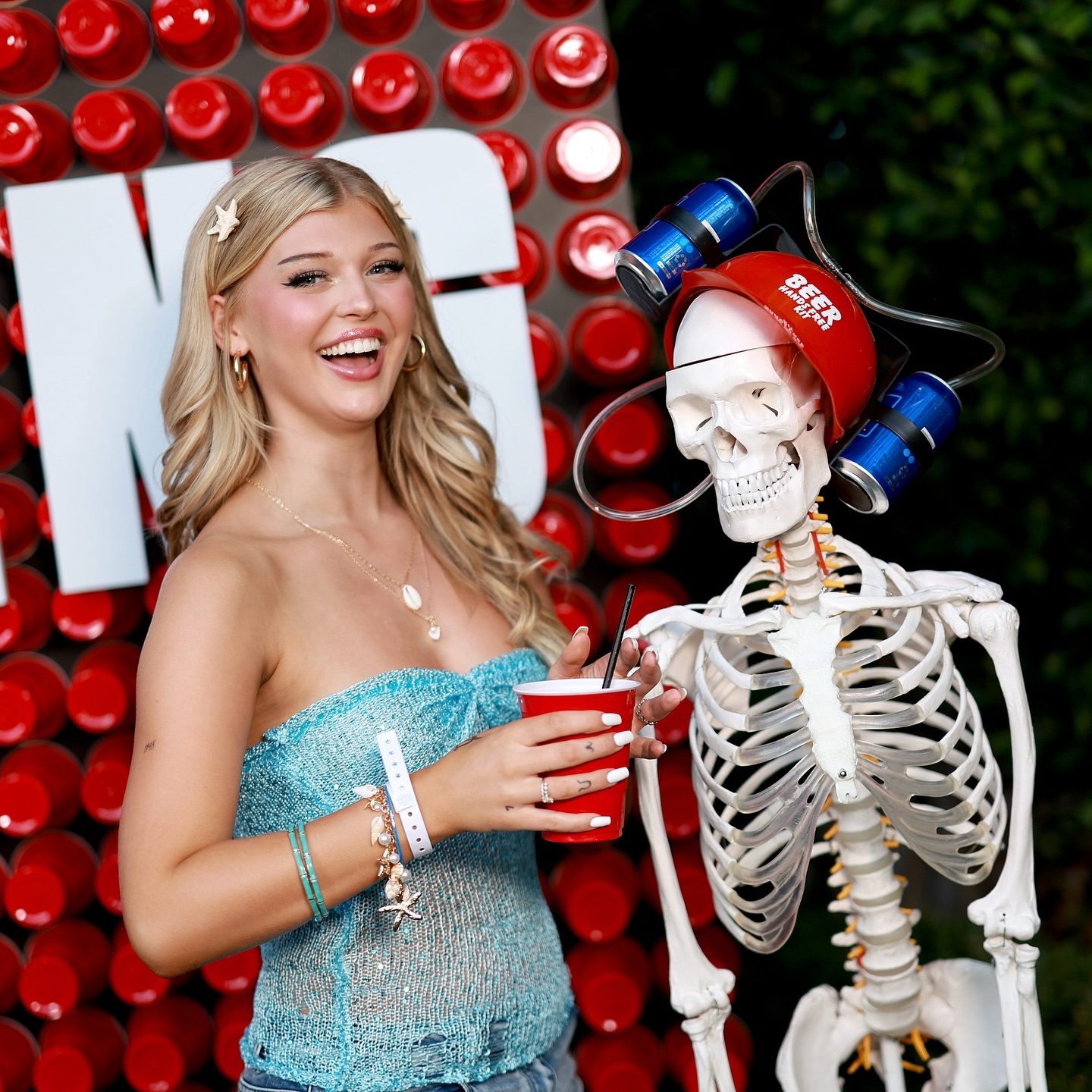 a smiling blonde woman in a blue strapless top and jeans poses next to a skeleton wearing a red "beer basics" beer helmet with attached cans and tubes. she holds a red cup with a black straw. the background shows a red and white decorative wall.