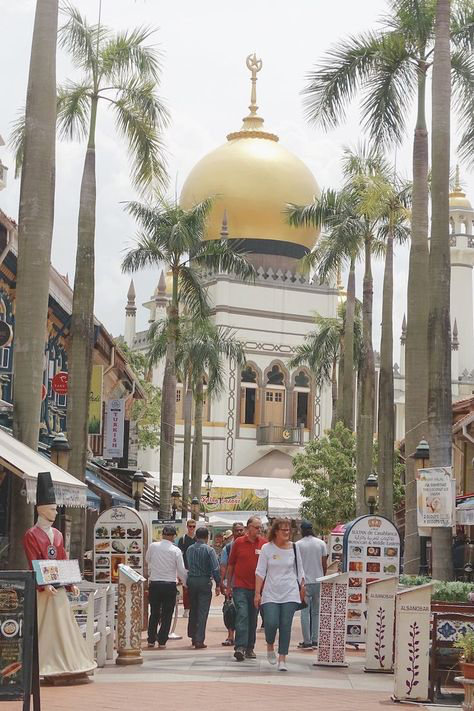 the image shows a vibrant street scene in singapore, with the iconic sultan mosque in the background. the mosque's golden dome is prominent, framed by tall palm trees. the foreground showcases a pedestrian walkway lined with shops and food stalls, displaying colorful signage and traditional designs. tourists and locals are seen strolling along the street, creating a lively atmosphere.  the overall impression is one of a bustling, culturally rich area blending modern and traditional elements.