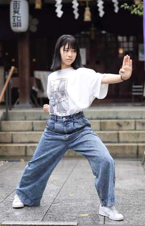 a young woman with dark hair in a white graphic tee and widelegged jeans strikes a confident martial arts pose in front of a japanese temple. the steps and traditional architecture of the temple provide a serene backdrop to her powerful stance. her outfit blends casual street style with the energy of her movement. the natural daylight illuminates the scene, highlighting the textures of her clothing and the details of the temple behind her.