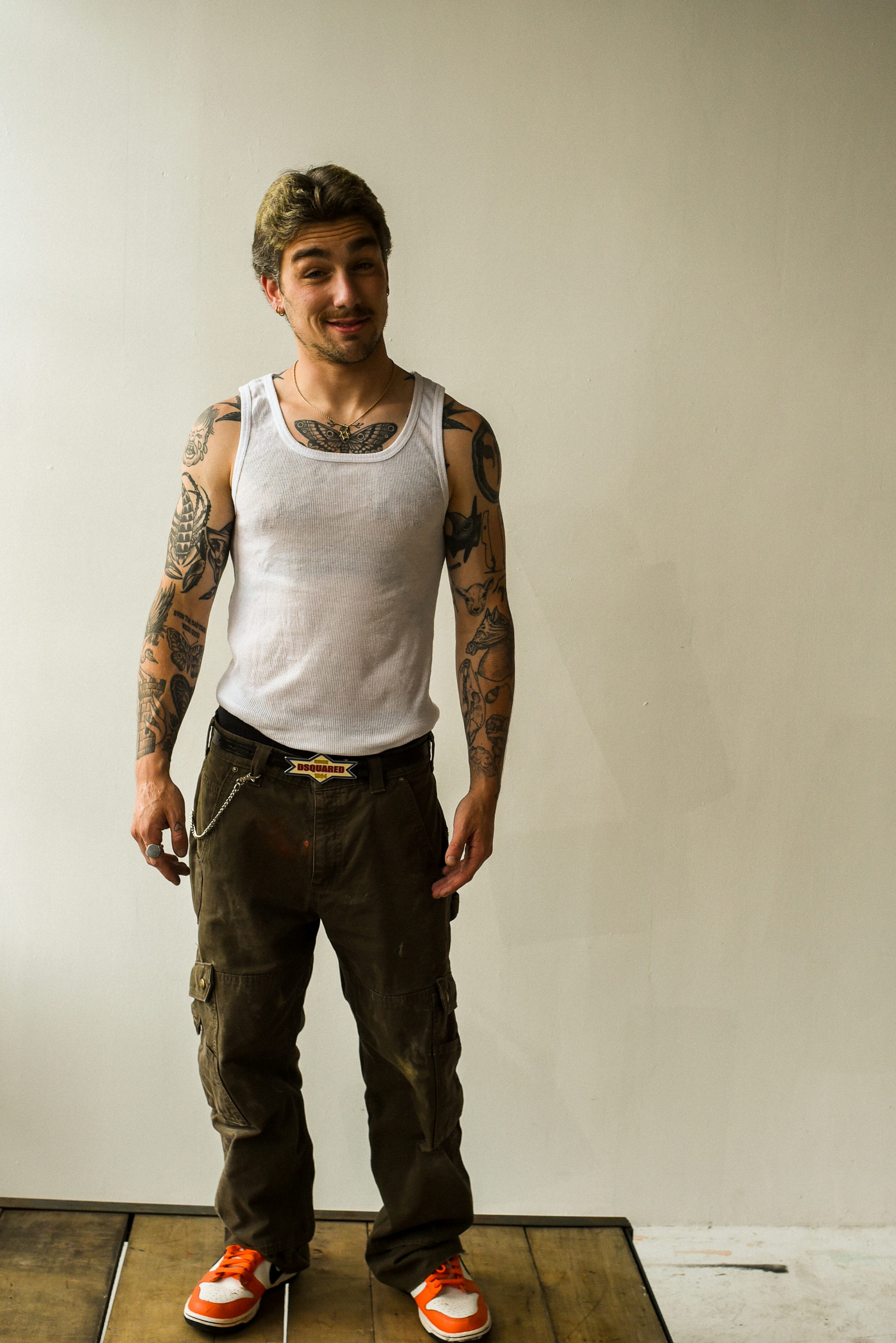 a man stands on a wooden platform, wearing a white tank top, brown cargo pants, and orange and white sneakers. he has many tattoos on his arms and chest, and wears a belt with a decorative buckle and a silver necklace. he has brown hair and is smirking slightly. he is standing in front of a plain, offwhite wall.