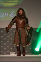 a man in a medieval warrior cosplay costume stands on a stage. he is wearing leather armor, a brown tunic, and green accents. he carries a sword and a shield. the background is dark with green lighting effects.