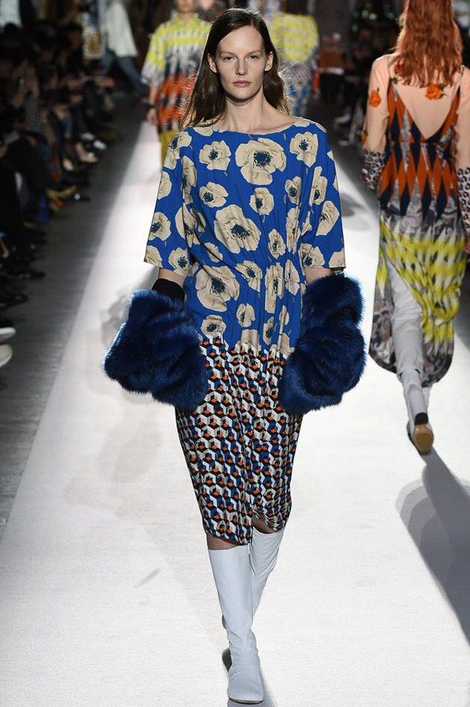 a fashion model walks down a runway wearing a unique dress with a floral top and a geometric bottom. the dress is blue and white with pops of orange and brown. she is also wearing long white boots and blue fur gloves. other models are visible in the background, showcasing a similar style. the runway is white and the overall setting suggests a professional fashion show.