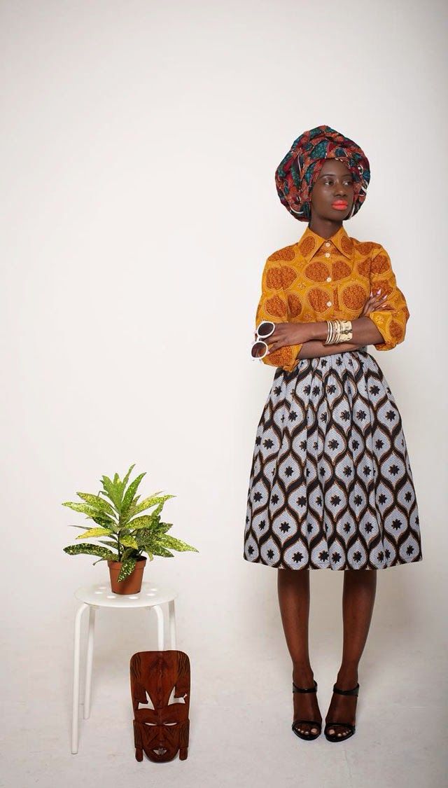 a stylish darkskinned woman is standing in a studio shot against a white background. she is wearing a yellow patterned blouse, a black and white patterned midi skirt, a colorful headwrap, sunglasses, and gold bangles.  she is also wearing black heeled sandals.  her arms are crossed across her chest. a small plant in a pot sits on a white stool next to her; a wooden mask is placed in front of the stool. the overall aesthetic is one of contemporary african fashion.