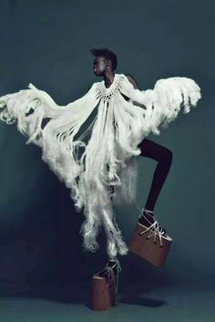 a model in a white, feathery dress with platform shoes poses against a green background.  she is wearing a white, feathery dress that is flowing and dramatic.  the model is looking off to the side, and her pose is very graceful.  the background is a simple bluegreen color, which allows the model's outfit to stand out.