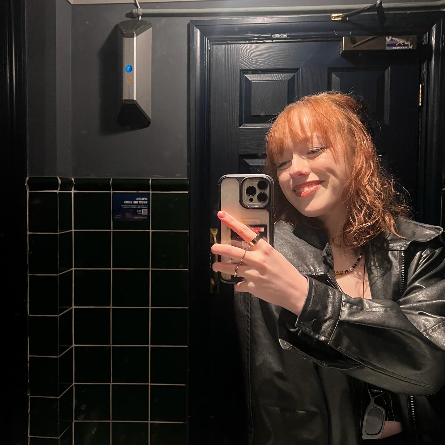 a young woman with red hair takes a selfie in a dark restroom. she's wearing a black leather jacket and several rings. the bathroom has black tile walls and a dark door in the background.  a hand sanitizer dispenser is visible on the wall. the overall mood is dark and moody.