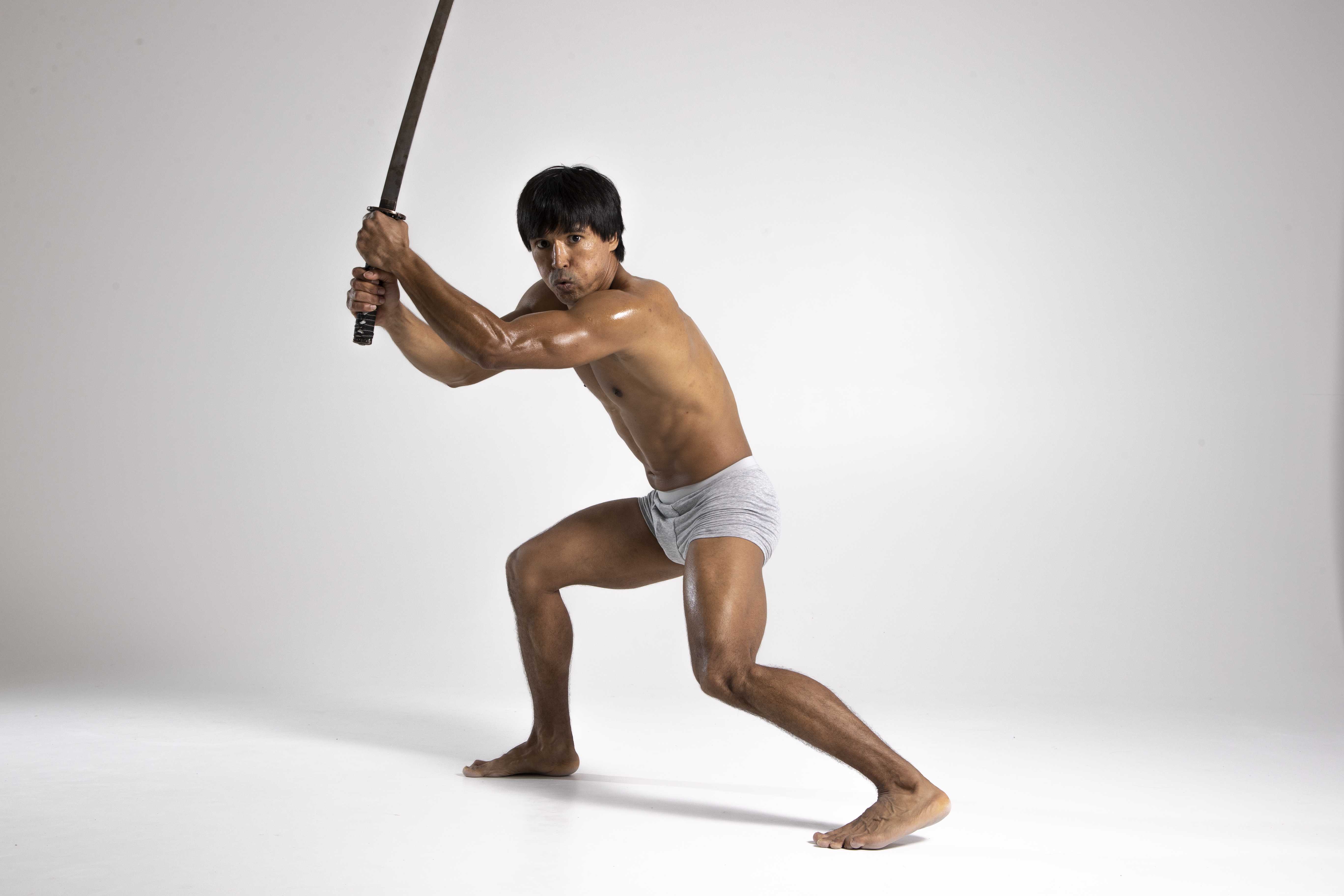 a shirtless man in underwear strikes a dramatic pose with a sword in a studio shot. he is barefoot and appears to be practicing martial arts.