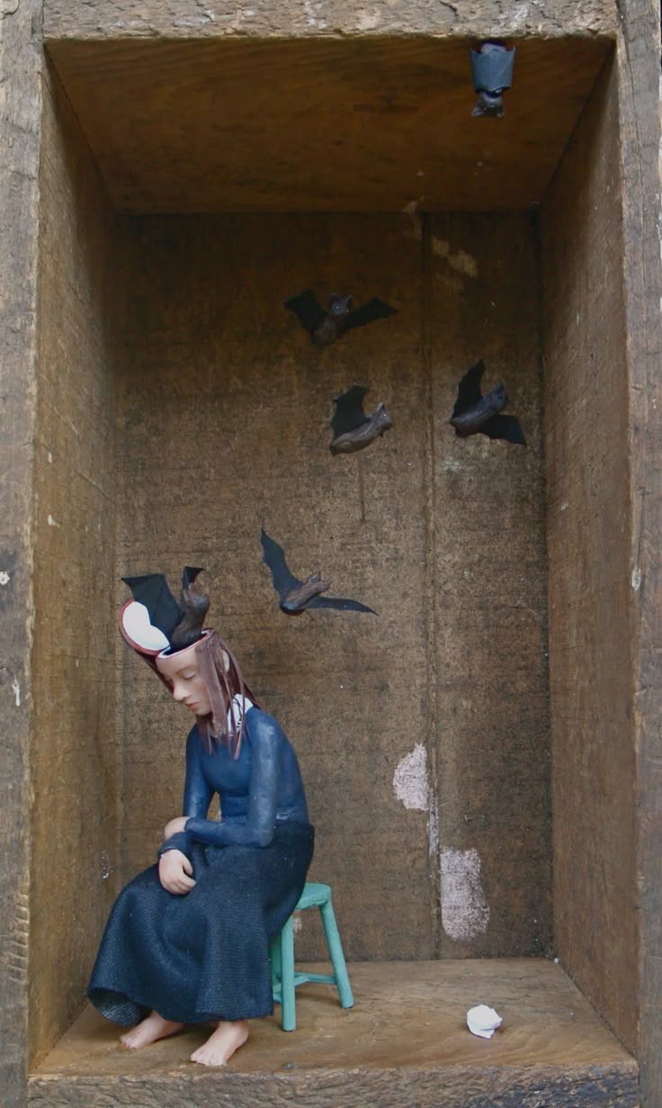 a photograph of a sculpted art piece depicting a melancholic figure seated on a stool inside a wooden boxlike structure. the figure is a young woman with an open head from which bats are emerging, other bats are depicted hanging on the walls and flying around her. a crumpled piece of paper lies on the floor. the overall mood is surreal and introspective.