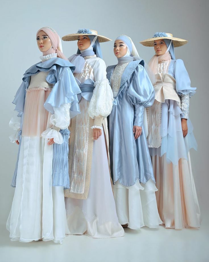 the image showcases four models, each wearing a unique, elegantly designed long dress and hijab in pastel shades of light blue and beige. the dresses are layered and feature flowing fabrics, giving them a dreamy, ethereal quality. each model also sports a widebrimmed hat, complementing their outfits. the overall aesthetic is high fashion and sophisticated, with a possible bridal or spring/summer influence. the backdrop is a simple, light grey studio setting, allowing the clothing to be the focal point. the photograph is professionally done, likely for a fashion magazine or website.