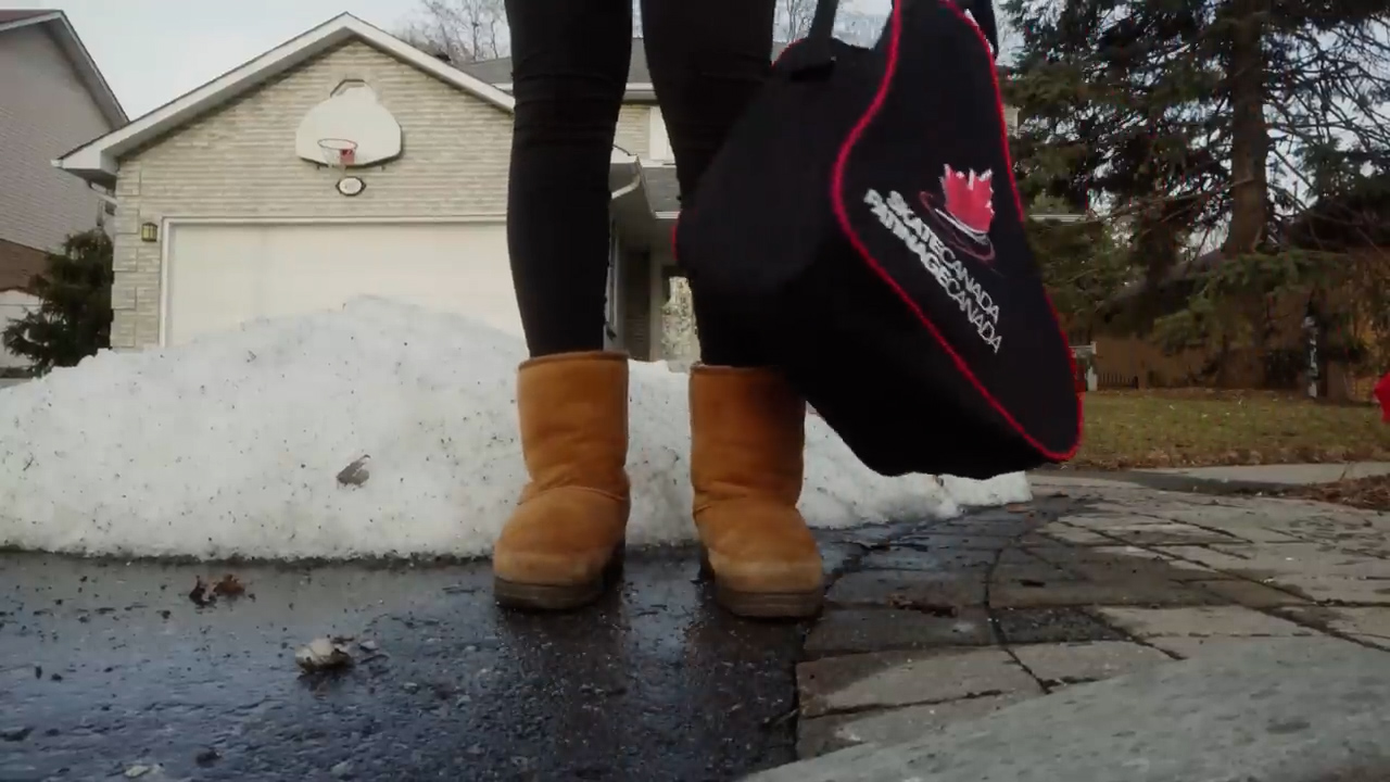 a woman is standing on a wet driveway with melting snow on the sides. she is wearing brown boots and black pants, and is holding a black sports bag. there is a basketball hoop above the garage door in the background. the image is taken from a low angle view.