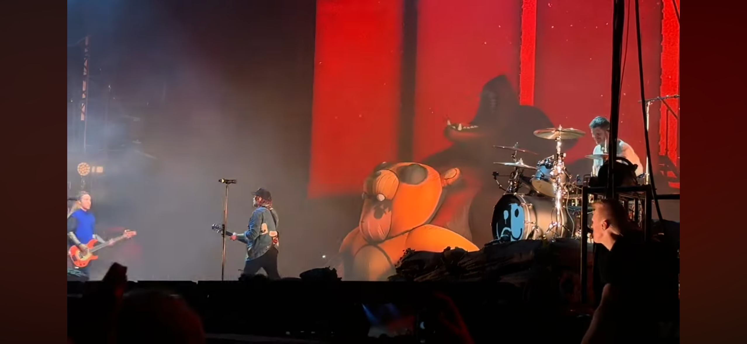 a concert stage with a band performing live. the stage is dimly lit with fog or smoke, and the band is playing in front of a large crowd. the band members are playing guitars, drums and bass guitar. the drummer is wearing a black shirt and is hitting the drums with passion. the guitarist is playing a  red guitar with a lot of energy. the lead vocalist is wearing a blue shirt and is singing into a microphone. the audience is clapping and singing along to the music, and the energy is high. there's a large screen in the background, showing a cartoon image of a bear.