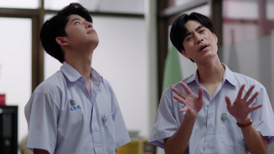 the image shows two young men wearing school uniforms, suggesting a scene from a thai bl (boys love) drama. they are standing indoors, possibly in a classroom or a school hallway, bathed in bright, natural light that enters from a window out of frame. the man on the left looks upwards, while the one on the right gestures with his hands while looking up and slightly to the right. both have a hopeful expression on their faces.