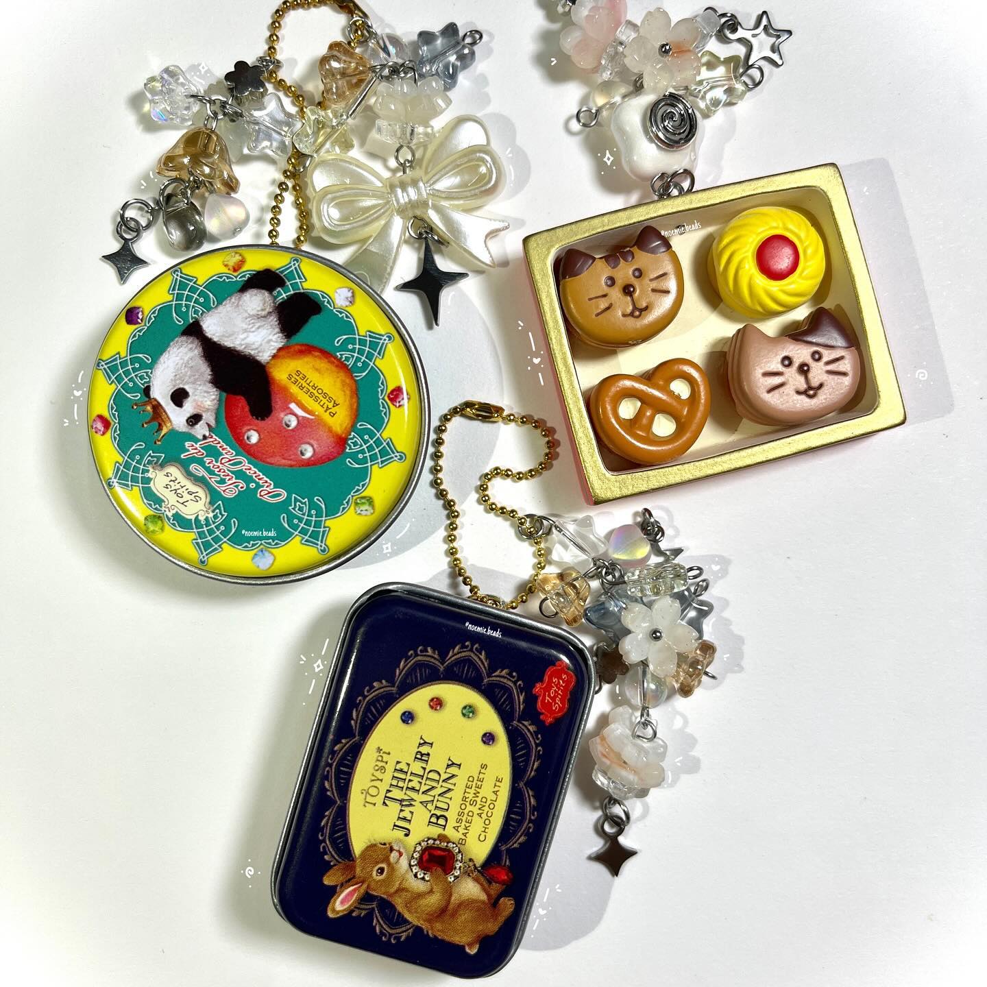 the image contains three different tins, two of which have charms hanging from them, and a box of sweets. the first tin is round with a yellow border and a panda sitting on a red ball in the middle. it has a gold chain and several charms hanging from it, including a bow and stars. the second tin is rectangular, dark blue, and has a picture of a bunny on it. it also has a gold chain and several charms hanging from it, including flowers and stars. the box of sweets contains four different treats: two catshaped sweets, a pretzel, and a yellow sweet with a red dot.