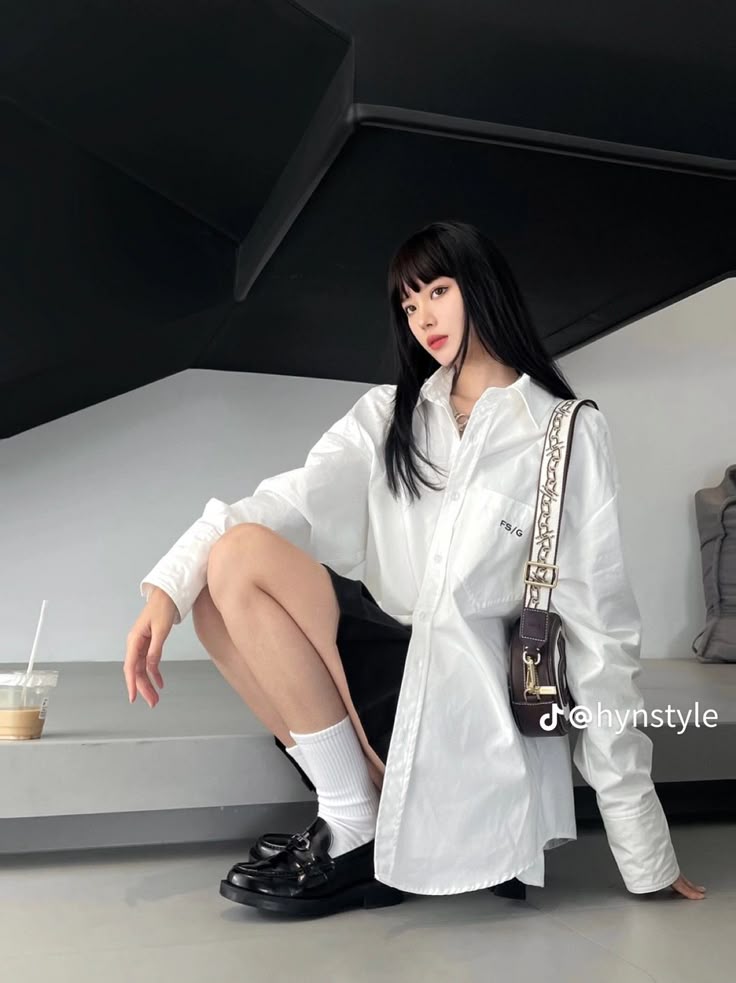 a woman is squatting on a bench. she is wearing a white shirt, black shorts, socks, and black shoes. she has a bag on her shoulder. she has bangs and long hair. there is a cup next to her. the background is black and white.