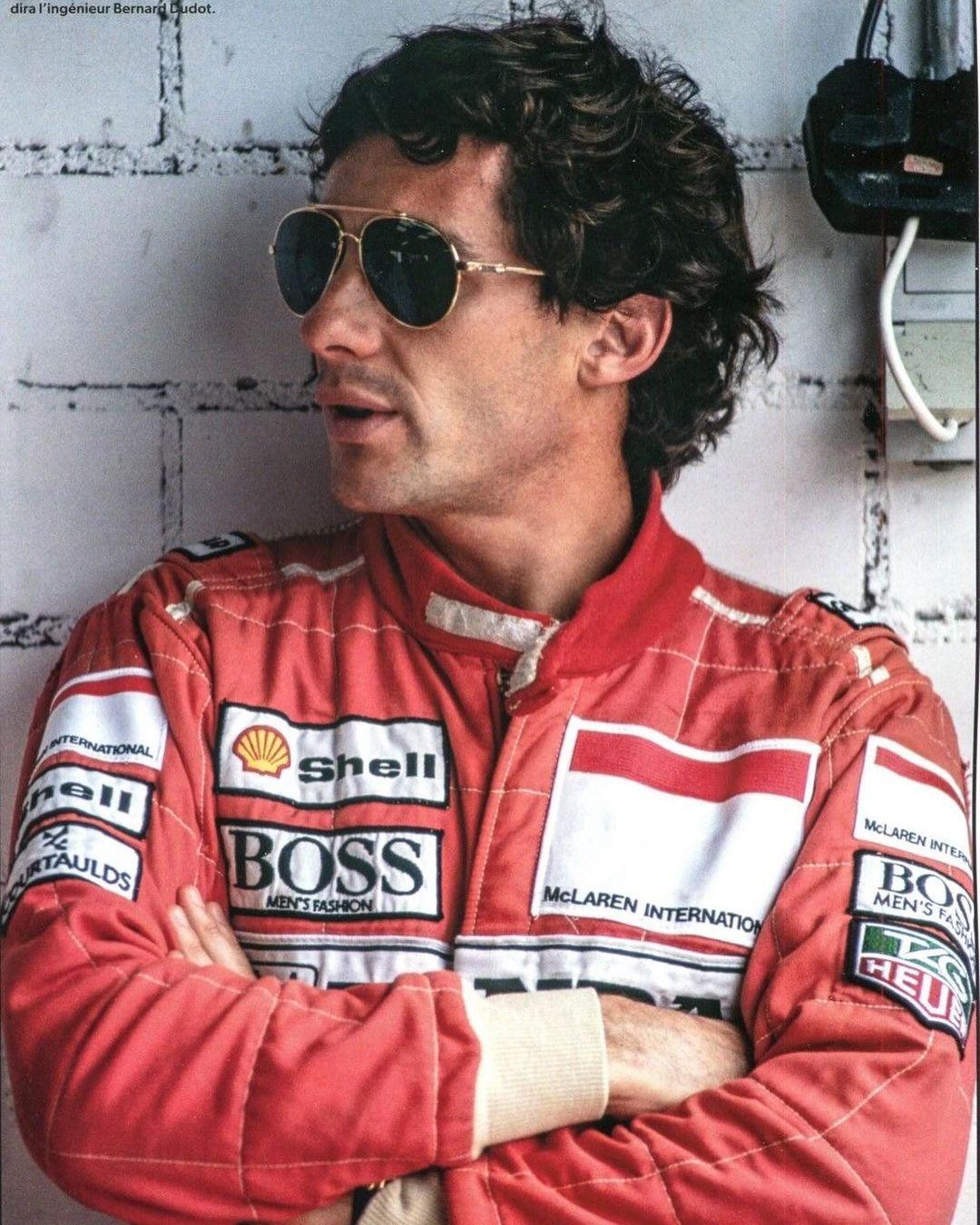 a race car driver in a red, white and black racing jacket with the logos of shell, boss, mclaren and heuer. he is wearing sunglasses and is looking to the left of the camera. he is standing in a garage.