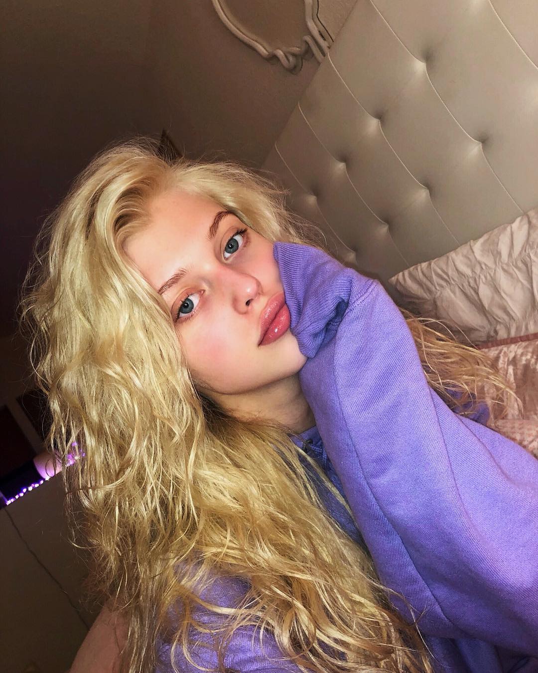 a young woman with blonde hair and blue eyes is lying down on a bed, wearing a purple hoodie. she is looking at the camera, her face resting on her hand. the background includes a headboard and parts of the bedroom. it appears to be a selfie taken indoors.