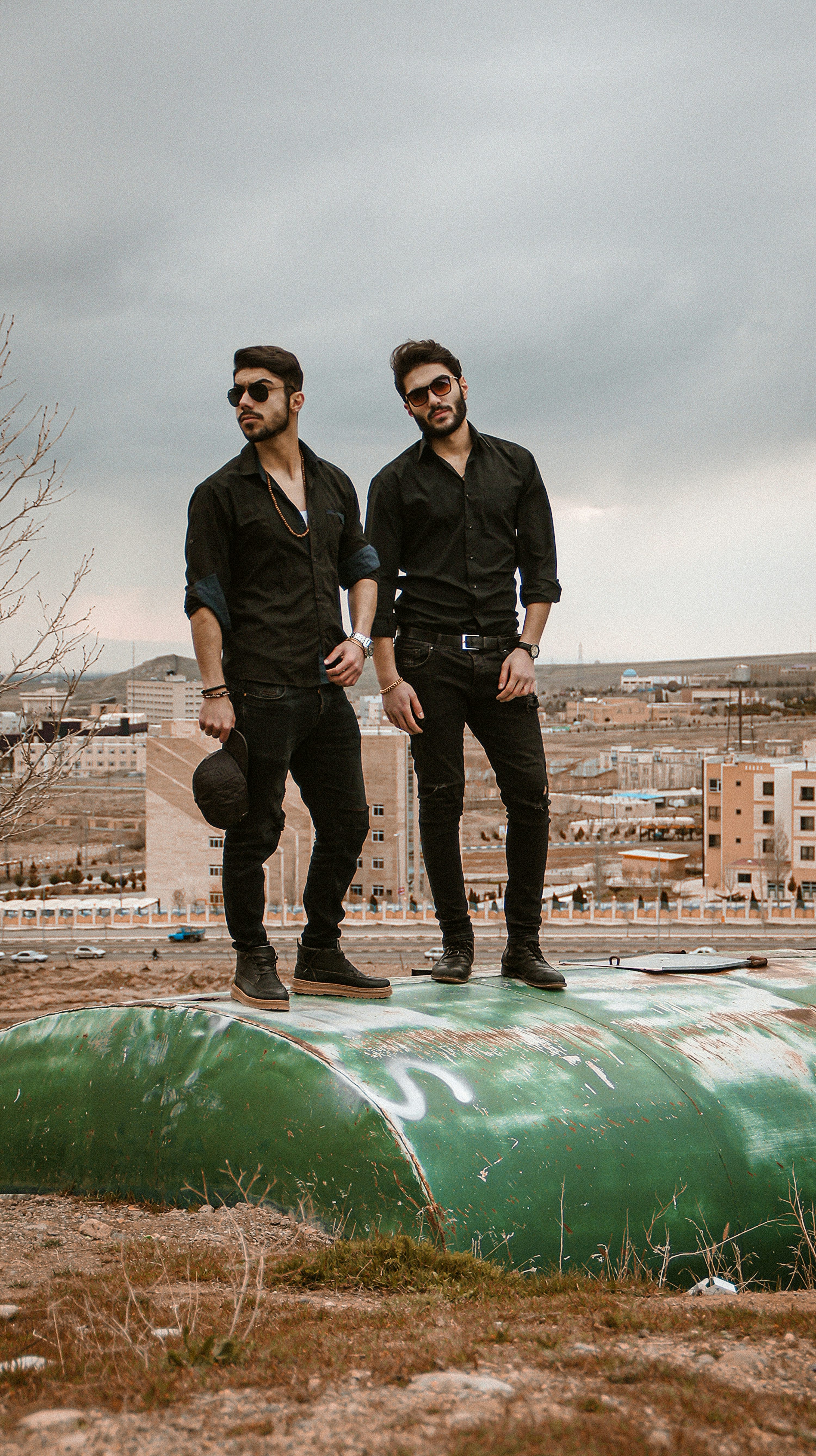 the image shows two identical twin brothers standing on a weathered green metal structure atop a hill overlooking a cityscape. they are dressed in similar outfits: black longsleeved shirts, black pants, and sunglasses.  they appear to be in their twenties and have a stylish, somewhat grunge aesthetic.  the background features a hazy, overcast sky and a mix of urban and undeveloped land, creating a contrast between the twins' sharp look and the rougher surroundings. the overall mood is stylish and slightly melancholic, reflecting the urban landscape's subdued colors and the twins' somewhat serious poses.