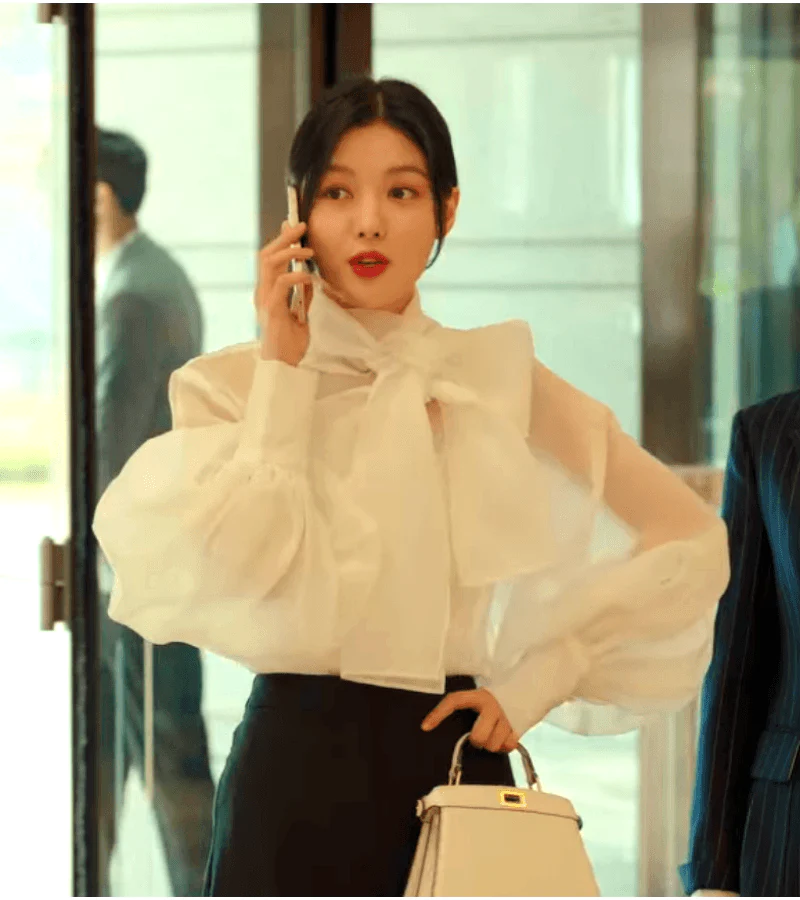 woman dressed in a stylish white blouse with a large bow and black pants stands in a hotel lobby, holding a phone to her ear and a handbag in her other hand. a man in a suit is blurred in the background. the scene is brightly lit with artificial light and captured from an eyelevel perspective.