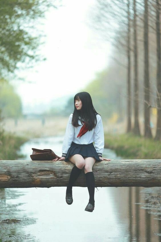 young woman in a school uniform, possibly japanese, sits on a large fallen log spanning a creek in a forest. she is turned slightly to the left, gazing into the distance with a contemplative expression. the environment is serene and peaceful, with soft, diffused light filtering through the trees. the overall color palette is cool and muted, emphasizing the natural beauty of the scene.