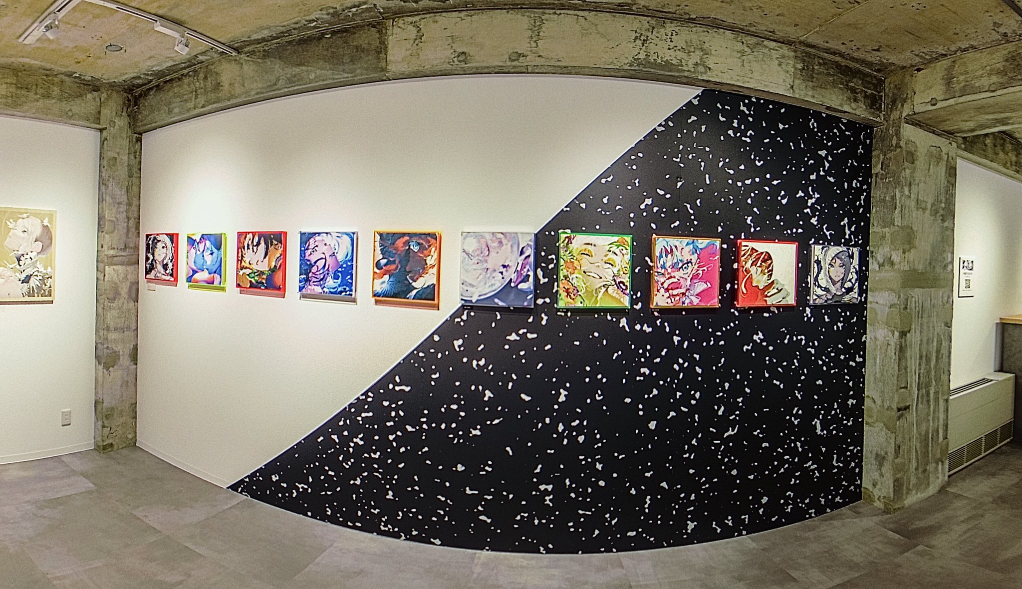 this wide shot shows the interior of an art gallery with a series of colorful, square, graphic paintings hung in a row on a white wall.  the far wall features a dramatic design with one half painted white and the other half covered in black wallpaper with white speckles. the concrete ceiling and the tiled floor suggest an industrial chic aesthetic.