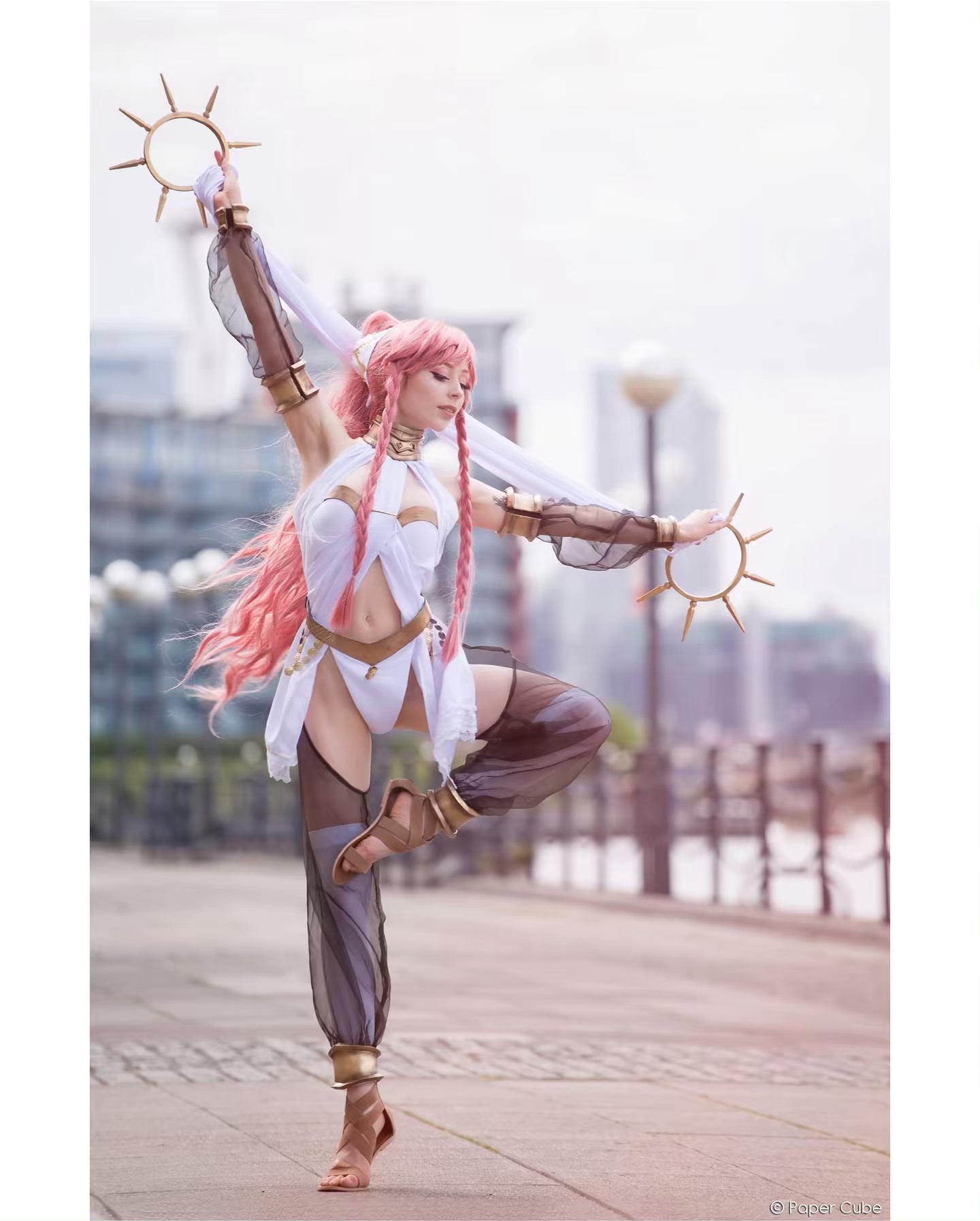 a full shot of a woman cosplaying a fantasy character. she has long pink hair, is wearing a white and gold costume with sheer black pants and sandals. she is posed elegantly on one leg holding two gold sunlike props. the backdrop is a city street.