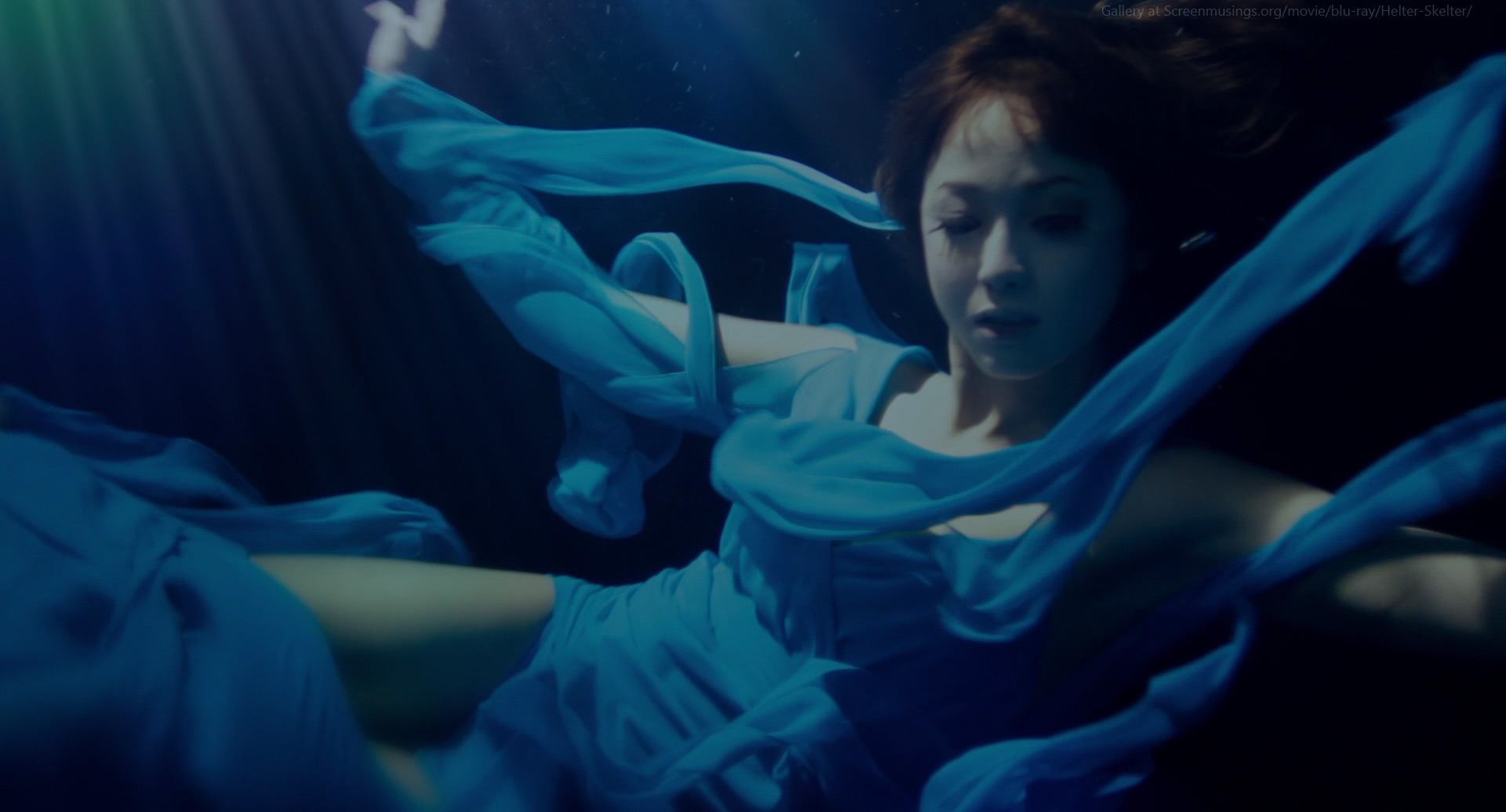 woman in a flowing blue dress is submerged underwater. she appears to be floating gracefully, her dress billowing around her. beams of light penetrate the water from above, creating a dramatic and ethereal atmosphere. the overall palette is predominantly blue, with shades ranging from deep navy to a soft aqua.