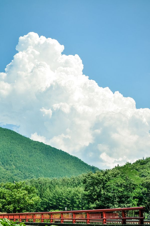 the image shows a large, billowing white cloud dominating the sky above a green landscape. a red bridge spans across the bottom of the image, with a densely forested mountain in the background, completing the scenic view under a bright blue sky.