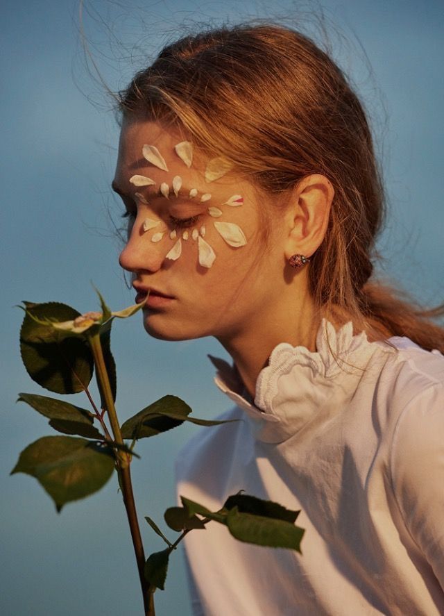 closeup portrait of a young woman with flower petals delicately arranged around her eye. she is wearing a white highnecked shirt with a ruffled collar and has long light brown hair pulled back in a loose ponytail. she is gently smelling a rose. the overall mood is serene, peaceful and dreamy. the lighting is soft and golden, creating a warm and romantic atmosphere. the image is aesthetically pleasing, featuring soft textures, delicate details, and natural elements.
