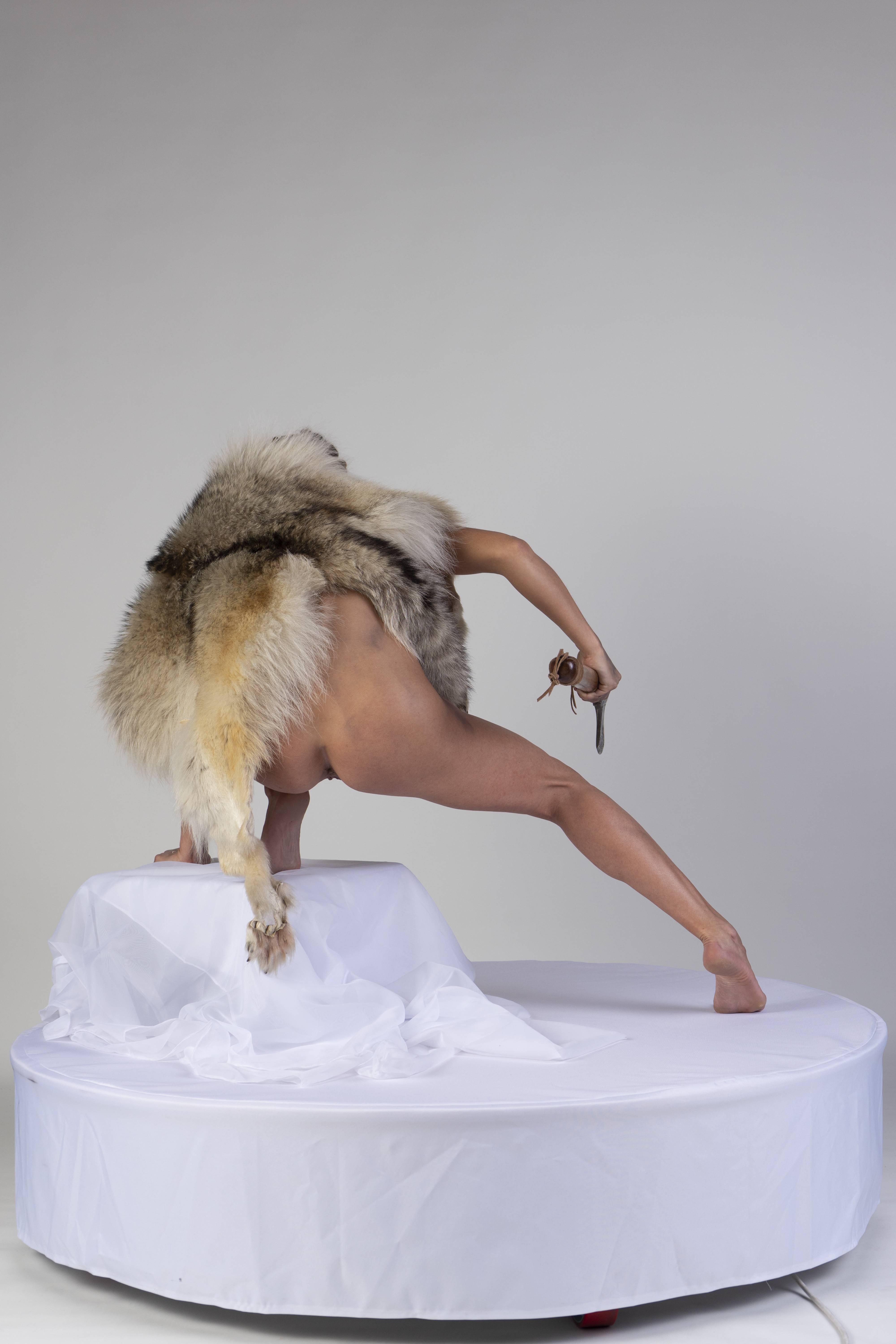 a nude person wearing fur clothing and holding a knife is standing on a platform indoors. the person is standing on a white platform in a brightly lit room. the person is mostly facing away from the camera, but their head is turned slightly to the left. the fur clothing is a large, shaggy piece of fur that covers most of the person's body. the person is holding a knife in their right hand. the knife is small and has a wooden handle. the background is a plain white wall.