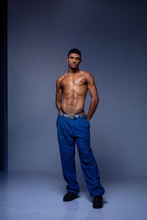 a shirtless man with an athletic build wearing blue pants and black shoes stands with his hands in his pockets in a studio setting. he is looking directly at the camera.