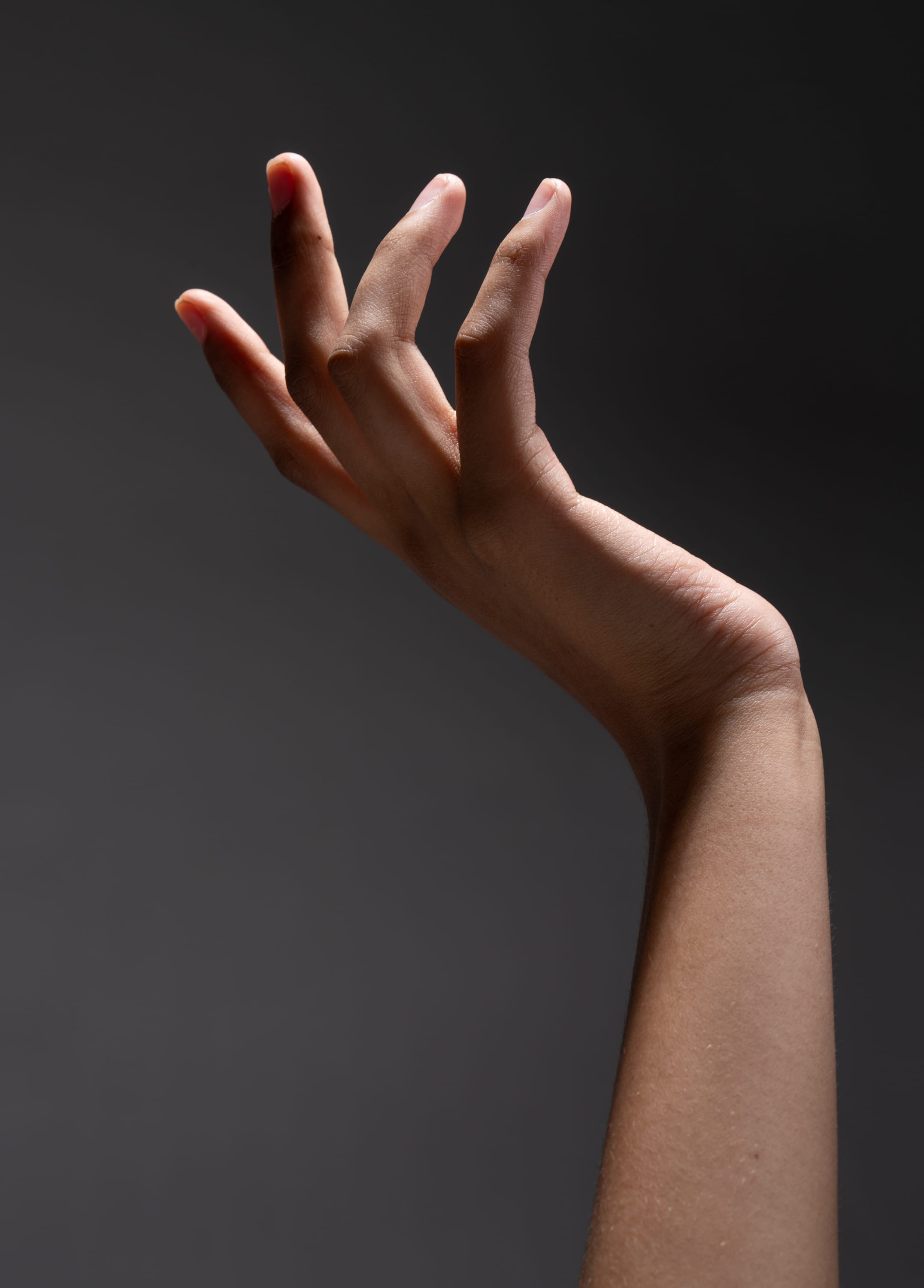 a woman's hand and arm are shown in a dimly lit environment. the hand is extended towards the left side of the image with the palm facing forward and the fingers slightly spread. the fingers are long and slender, and the skin is smooth and clear. the hand is illuminated from the front, creating shadows on the back of the hand and arm. the arm is extended upwards, with the elbow slightly bent. the background is a dark gray color, which contrasts with the lighter skin tones of the hand and arm. the image conveys a sense of elegance and grace.