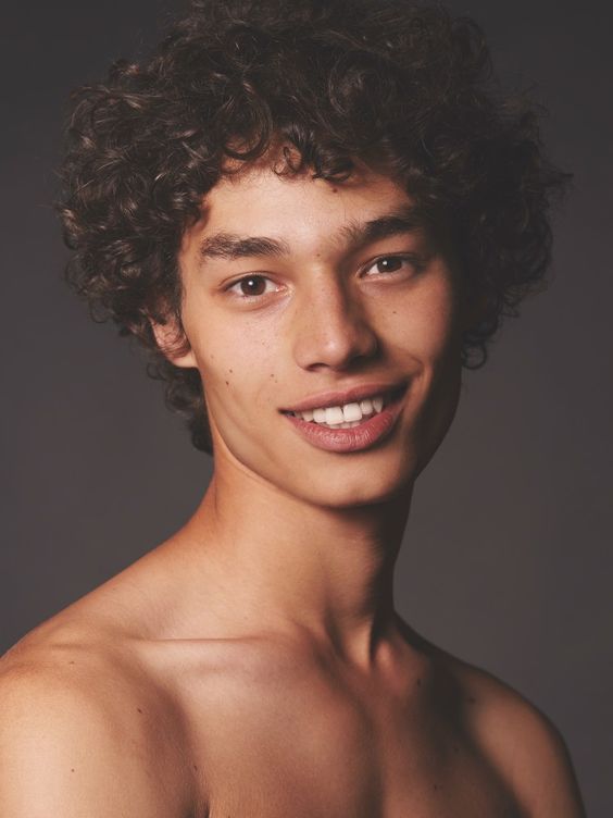 a headshot of a young adult male model with curly hair. he has bare shoulders and is smiling at the camera.  the background is dark grey.