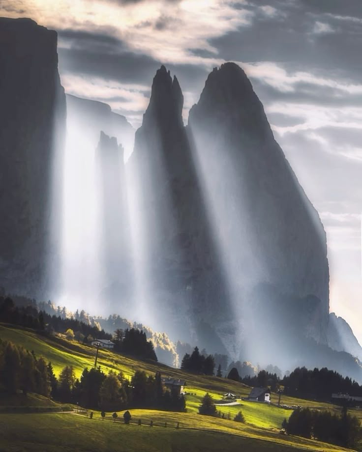 the image shows a dramatic landscape with tall, rocky mountains rising from a green valley. sunbeams cut through the atmosphere, creating a misty, ethereal effect. the valley is dotted with trees and small buildings. the sky is cloudy, adding to the overall moodiness of the scene.