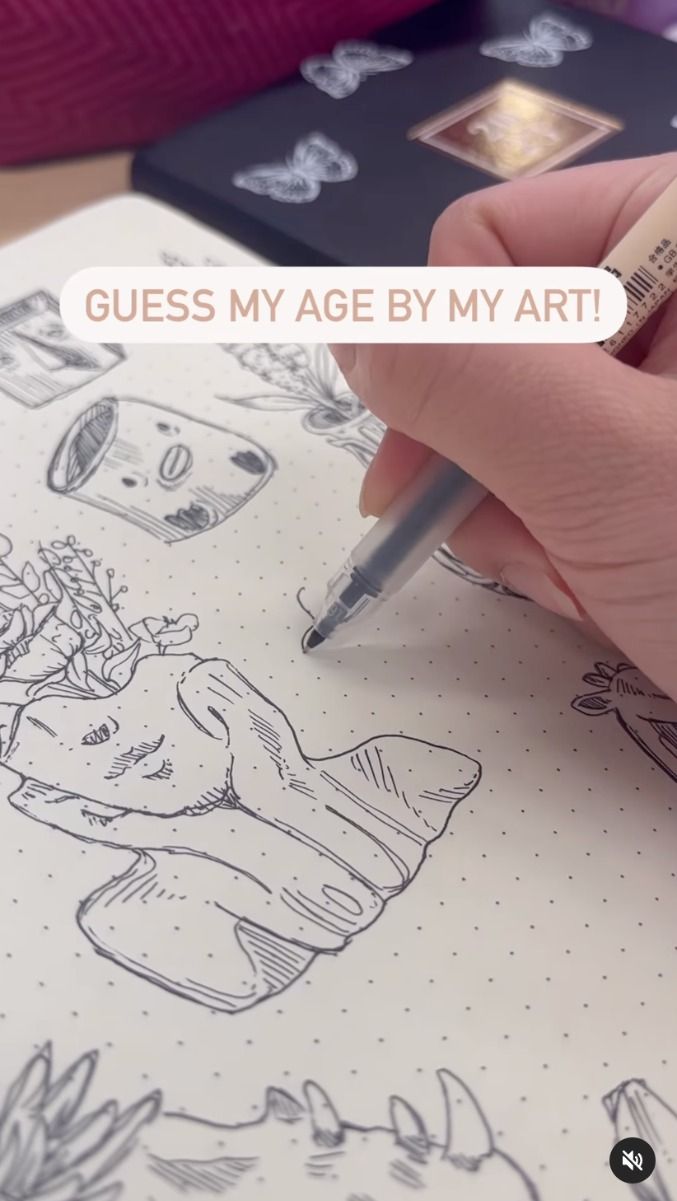 a closeup shot of a hand holding a fine liner pen, drawing intricate line art in a sketchbook. the art features whimsical drawings of flowers in pots, along with other abstract shapes and textures. the text overlay on the image says "guess my age by my art!".  the background includes a closed black notebook and a pink bag.  the style of art is delicate and detailed, suggesting a young person's artistic work.