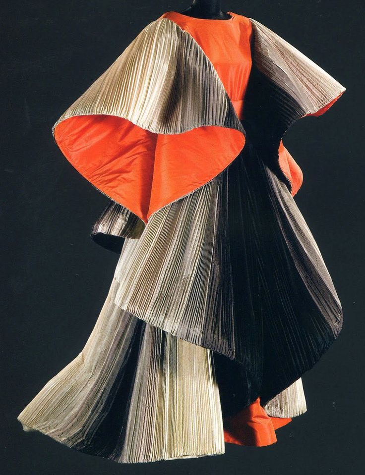 a unique and artistic dress on a mannequin against a dark background. the dress is constructed from pleated fabric in shades of white/beige and black, featuring an orange inner lining on the top and possibly throughout. the pleats create a textured and sculptural effect, and the design appears avantgarde and unconventional.