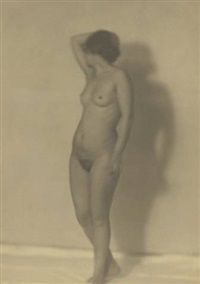 he image is a monochrome photograph of a nude female figure. the woman is standing in a slightly threequarter pose, her body oriented to the left, but her head turned slightly more towards the viewer. her left arm is raised, and her hand rests lightly on her head. her body is softly illuminated against a muted background, and her shadow is cast on the wall behind her. the overall style suggests a classical or vintage artistic approach, with a focus on form and a sense of tranquility. the image is softfocused, giving it an ethereal quality, and the low contrast and sepia tone contribute to the vintage feel.