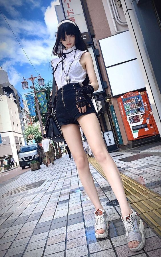 a young woman in stylish attire poses for a photo on a bustling japanese city street. the photo is taken from a low angle, emphasizing her height and the surrounding urban environment. the bright sunlight casts shadows and highlights the details of her outfit and the cityscape.