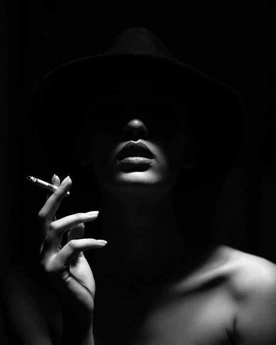 a woman wearing a hat, looks down,  and holds a cigarette between her fingers. she is lit in dramatic lighting, casting strong shadows around her face and body. she