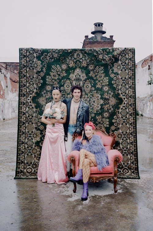 a group photo of three women in front of a large, ornate green carpet. two of the women are dressed in pink and purple, one in a long pink dress and the other in a purple fluffy coat and fishnet stockings. the third woman is shirtless and wearing a dark jacket. they stand in front of a dilapidated building in an outdoor setting. the atmosphere is grunge and alternative. the photo has a 90s aesthetic.